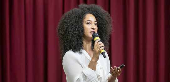 A fotografia mostra uma televisão e atrás um painel e uma cortina da cor vermelha. A tevê mostra...