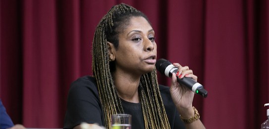 A fotografia mostra uma televisão e atrás um painel e uma cortina da cor vermelha. A tevê mostra...