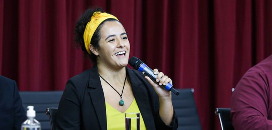 A fotografia mostra uma televisão e atrás um painel e uma cortina da cor vermelha. A tevê mostra...