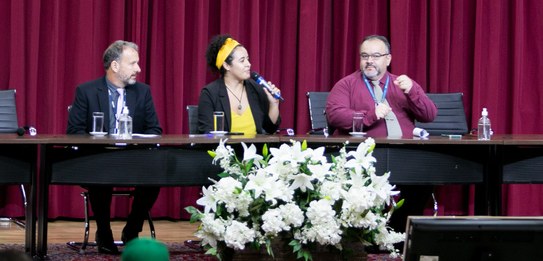 A fotografia mostra uma televisão e atrás um painel e uma cortina da cor vermelha. A tevê mostra...