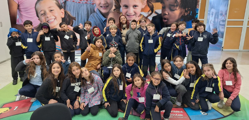 Fotografia de diversas crianças uniformizadas posando sorrindo em frente a um banner com a foto ...