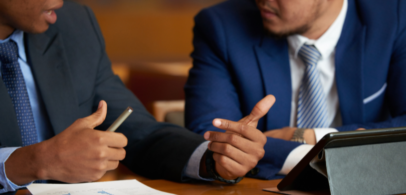 Fotografia em que se observa o tronco e as mãos de dois homens sentados à mesa em uma reunião de...