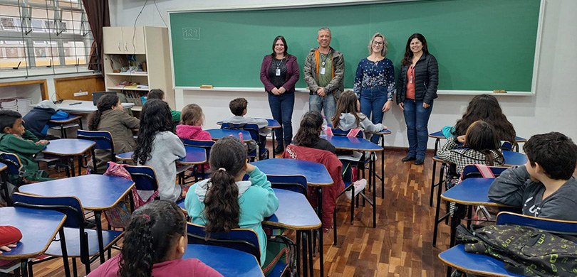 Fotografia de três mulheres e um homem falando para estudantes em uma sala de aula. Ao fundo, um...