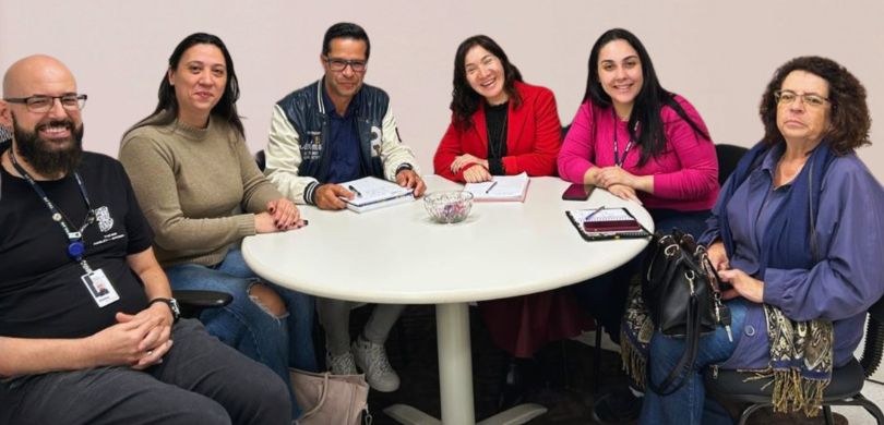 Fotografia de um grupo de seis pessoas, entre homens e mulheres, dispostas ao redor de uma mesa ...