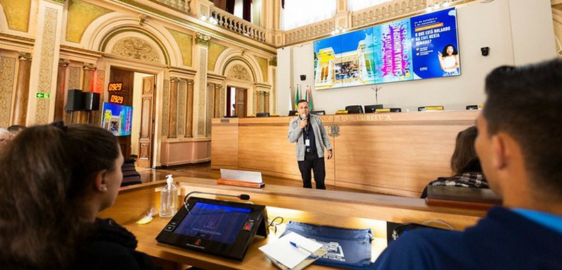 Fotografia de um homem falando em um microfone diante de algumas pessoas na Câmara Municipal de ...