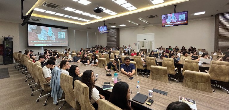 Sala ampla composta de duas grandes mesas retangulares, com cadeiras dos dois lados, quase compl...