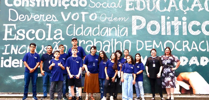 Fotografia de 15 pessoas, entre homens e mulheres, em frente a um banner de fundo verde, em que ...