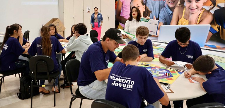 Fotografia de dois grupos de estudantes uniformizados reunidos ao redor de mesas redondas, enqua...