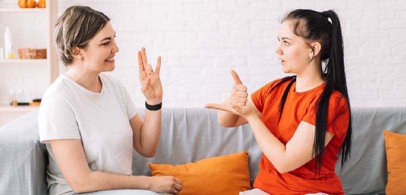 Na imagem, há duas mulheres conversando em Libras. Uma delas tem cabelos loiros, e veste camisa ...