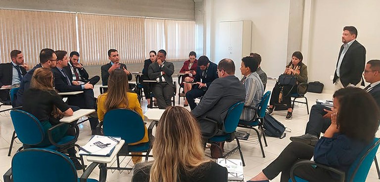Fotografia de diversas pessoas reunidas em um círculo em uma sala com paredes brancas. Elas estã...
