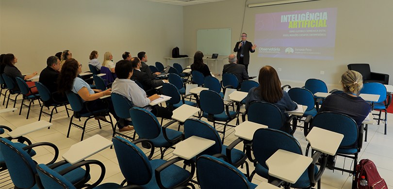 Fotografia de uma sala de aula onde um homem, vestindo terno e óculos, realiza uma apresentação ...