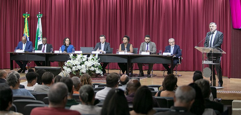 O local é um auditório. Na parte de baixo da foto há várias pessoas sentadas em cadeiras. No fun...