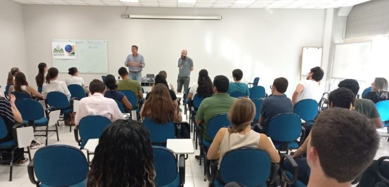 Fotografia em que aparecem vários estudantes sentados de costas em carteiras azuis. Ao fundo, do...