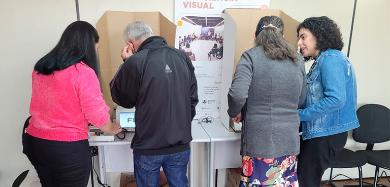 Fotografia de quatro pessoas em pé, viradas de costas. São três mulheres e um homem. Duas mulher...
