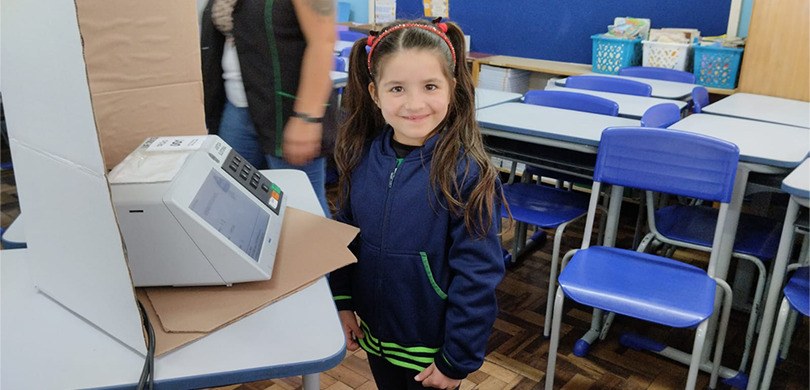 Fotografia de uma menina vestida com um uniforme escolar azul e verde sorrindo para foto em fren...
