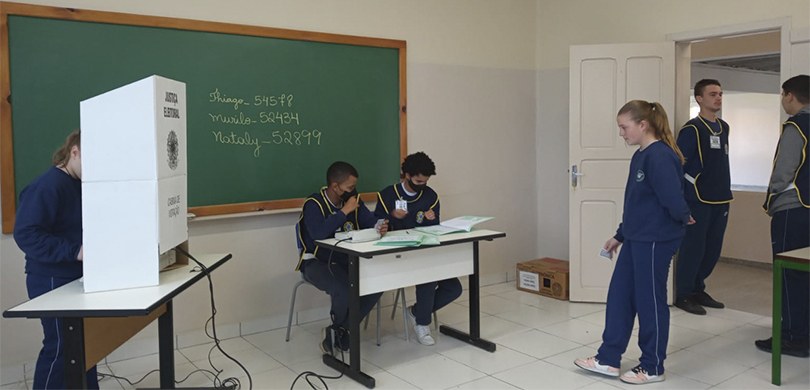 Fotografia de uma sala de aula, durante votação do Parlamento Jovem. Uma estudante está na cabin...