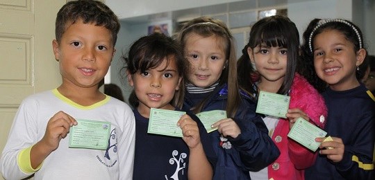 Fotografia de um grupo de crianças. Elas olham para a câmera e seguram um título de eleitor. 