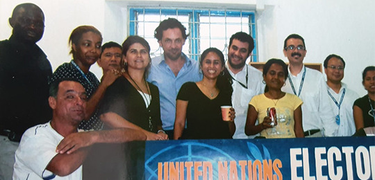 Em 2008, prestou serviço como conselheiro legal eleitoral, na missão da ONU no Timor Leste. Fotografia de um grupo de homens e mulheres. Atrás, há uma janela com grade. Na frente do grupo,...
