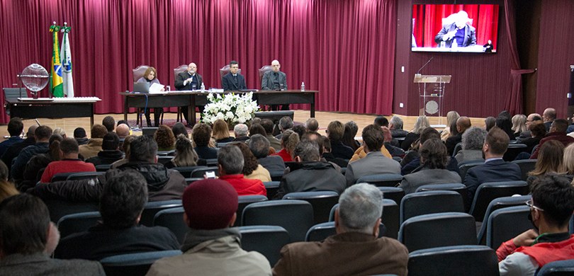 Fotografia de um auditório. Há pessoas sentadas na plateia e, no palco, uma mesa com quatro pess...