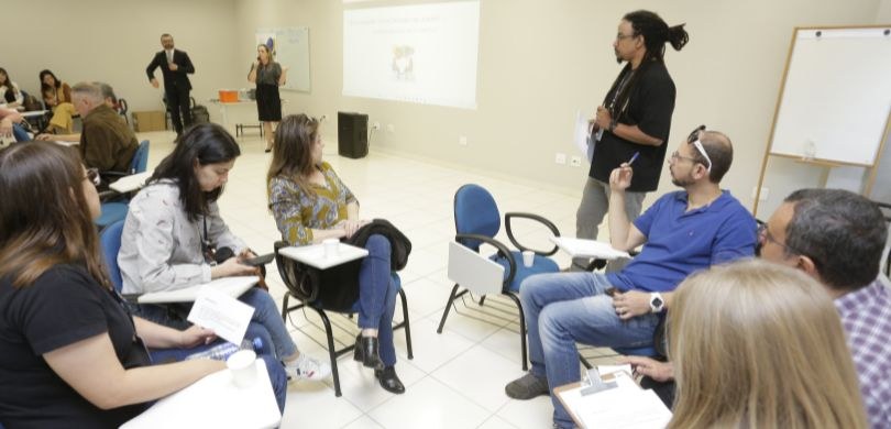 Fotografia de diversas pessoas, sentadas em carteiras escolares azuis, organizadas em formato de...