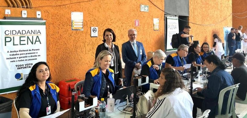 Fotografia de quatro mulheres e dois homens posando sorrindo. À frente, há mesas e cadeiras bran...