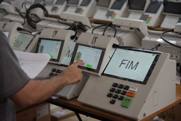 Fotografia de uma pessoa fazendo testes de votação em uma urna eletrônica. Várias urnas estão at...
