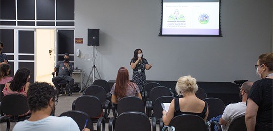 Fotografia de uma sala com pessoas assistindo a uma apresentação feita por uma mulher. Ao fundo,...