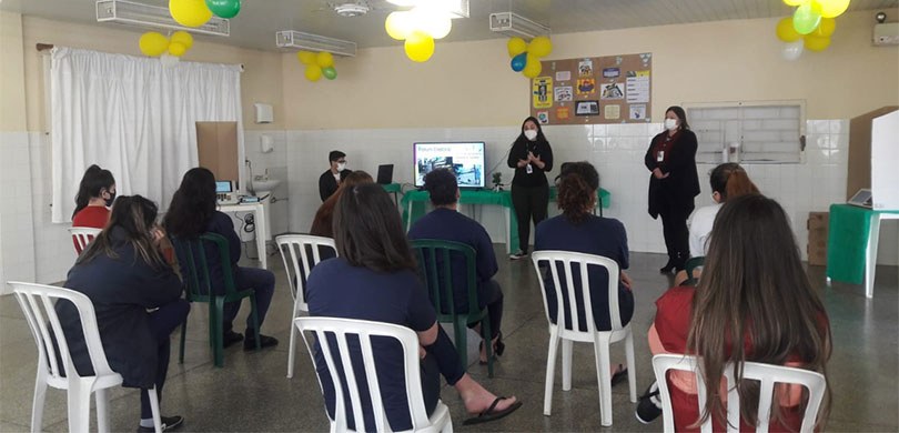 Fotografia de uma sala, com duas mulheres falando diante de uma plateia de nove mulheres. Uma ou...