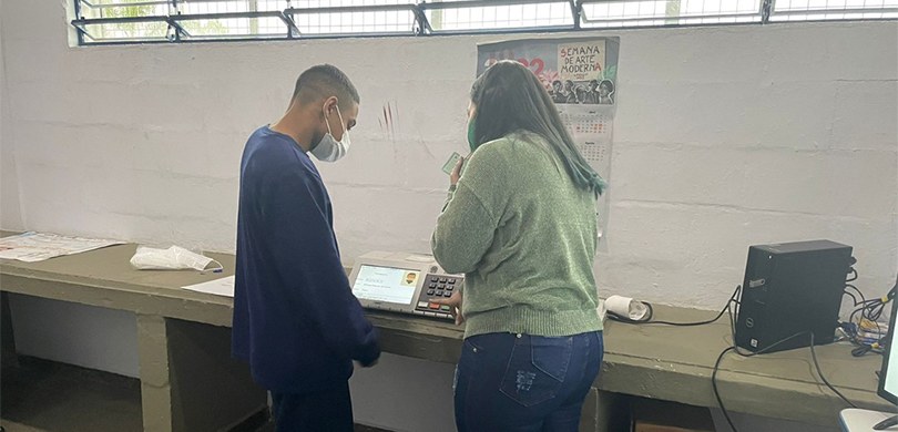 Fotografia de uma mulher apontando em direção à urna eletrônica enquanto um estudante a observa.