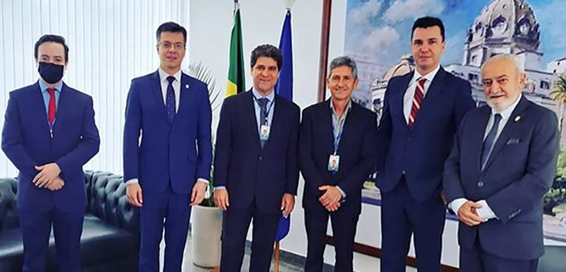 Fotografia de seis homens posando sorrindo para foto. Todos usam terno e gravata. Ao fundo, há u...