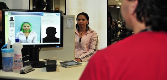 Foto de eleitora sendo atendida por funcionário da Justiça Eleitoral. À esquerda, uma tela de co...