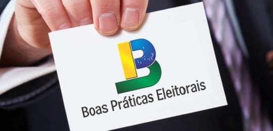 Foto de uma mão segurando um papel branco com a logo do projeto Boas Práticas (um "B" azul, verd...