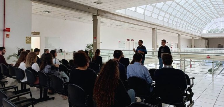 Fotografia de dois homens em pé palestrando para jovens, sentados em cadeiras pretas (longarinas...