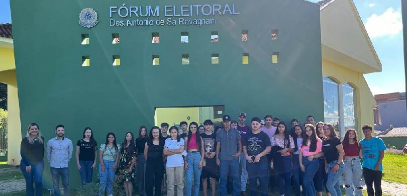 Cerca de quarenta jovens posam para a foto em frente a um prédio, com paredes verde e amarelas. ...