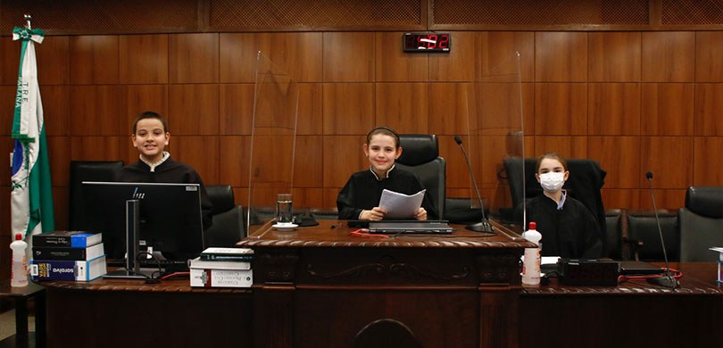 Fotografia de três crianças durante julgamento simulado na Sala de Sessões do TRE-PR. São duas m...