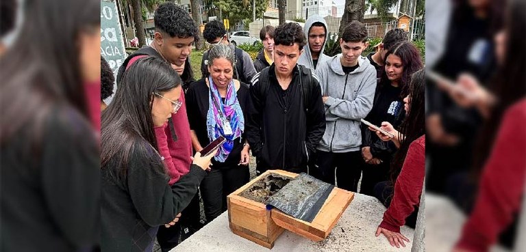 Estudantes do Colégio Estadual Iara Bergmann realizam alistamento em Curitiba 2 Vários estudantes estão em torno de uma bancada sobre a qual se vê uma caixa de madeira com uma ...