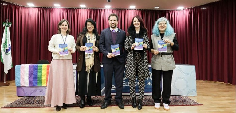 Fotografia de cinco pessoas posando sorrindo em um palco de auditório com uma cartilha em mãos. ...