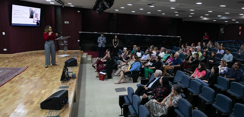 Fotografia do palco de um auditório, em que se vê uma mulher loira, com camisa vermelha e calça ...