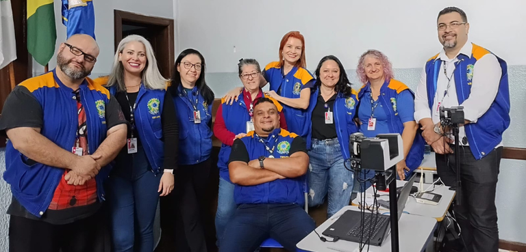 Fotografia de homens e mulheres posando sorrindo em uma sala com paredes branca e cinza. Eles ve...