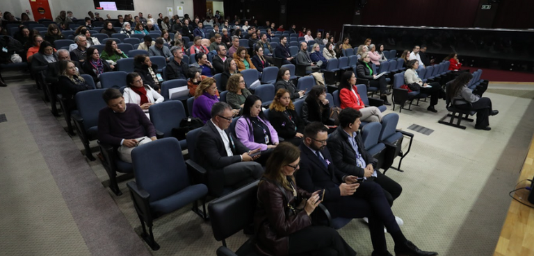 Fotografia em que se observam várias pessoas sentadas em poltronas azuis de um auditório. O piso...