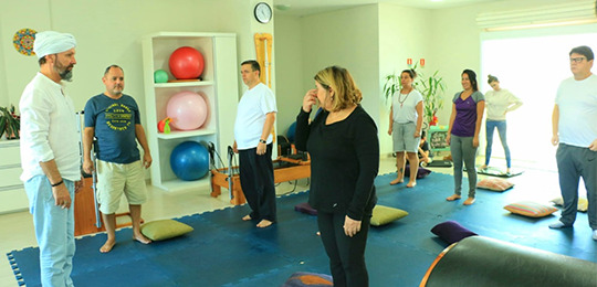Fernando é professor de yoga e yogaterapia Fotografia de um grupo de pessoas em pé em uma sala de atividades físicas. O chão é coberto por ...