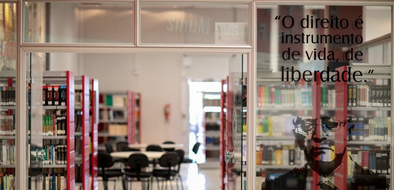 A foto é da entrada da biblioteca. Há janelas e uma porta de vidro. Atrás mostra várias estantes...