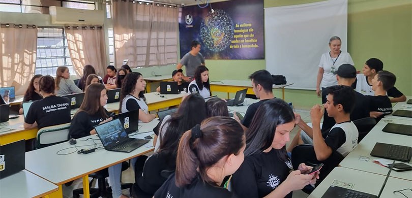 Fotografia de uma sala de informática em um colégio. Vários estudantes uniformizados estão prese...