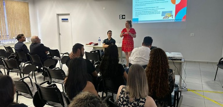 Fotografia em que se veem, ao fundo, de frente para a plateia sentada de costas, uma mulher em p...