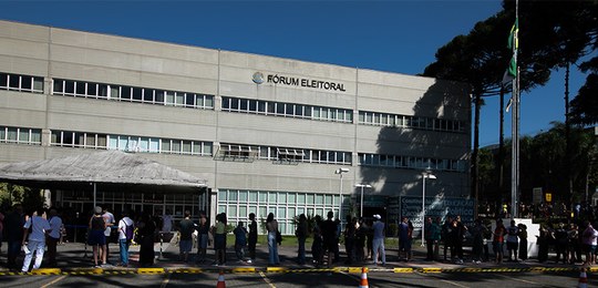 Foto do Fórum Eleitoral de Curitiba. Em primeiro plano, são vistas pessoas em uma fila. Ao fundo...