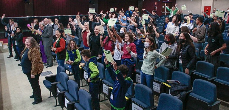 Fotografia das alunas e alunos no auditório do TRE-PR. Todos estão em pé participando da gravaçã...