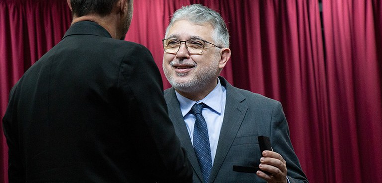 Fotografia de dois homens se cumprimentando. Um deles aparece de costas e o outro sorrindo. Fotografia de dois homens se cumprimentando. Um deles aparece de costas e o outro sorrindo.