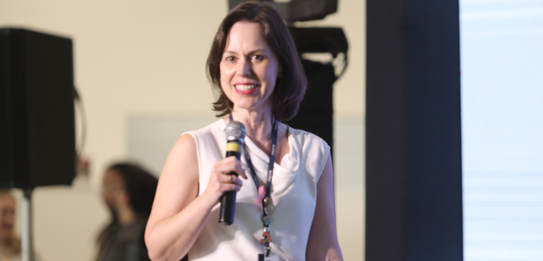 A Diretora-Geral Solange Maria Vieira fala para a plateia segurando um microfone. Atrás dela, um...