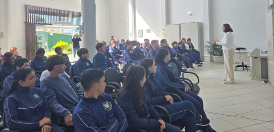 Fotografia de uma mulher em pé palestrando para jovens uniformizados em uma sala de aula com par...