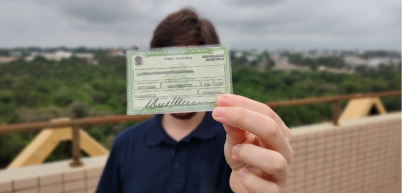 Fotografia de um homem jovem segurando um Título Eleitoral na frente do seu rosto. Ele tem cabel...
